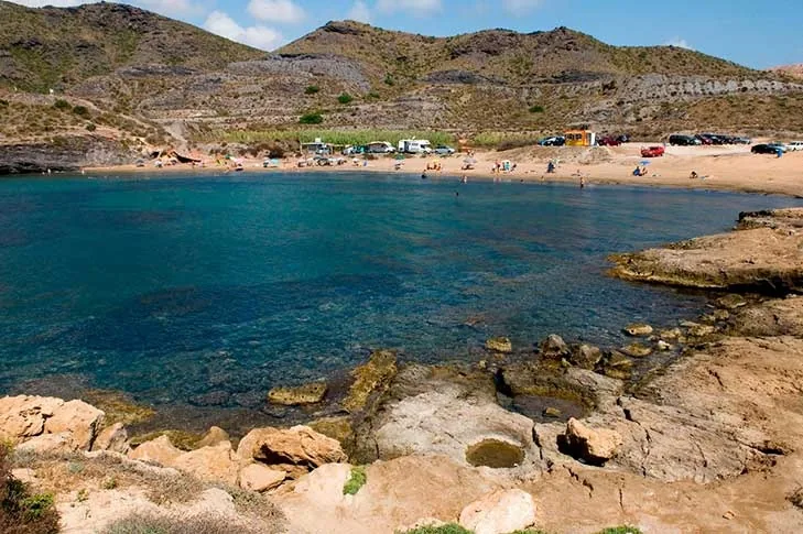 Cala Reona seen from above, with turquoise water, rocks in the foreground and a scrub-covered slope