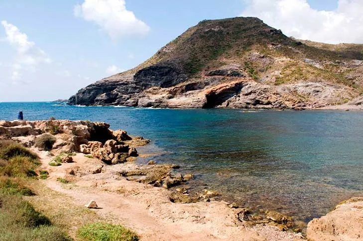 Cala Reona with the rocky hill behind, turquoise water and a dirt path through scrub