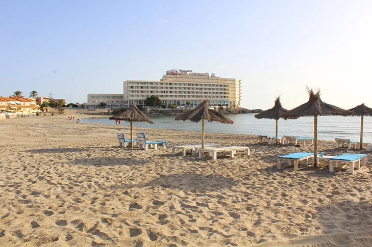 Parasols coniques en paille et transats bleus à Playa Galúa, avec l'hôtel de La Manga au fond