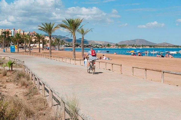 Holzsteg von Playa Paraíso mit einem Radfahrer, Sonnenschirmen und Badegästen im Sand