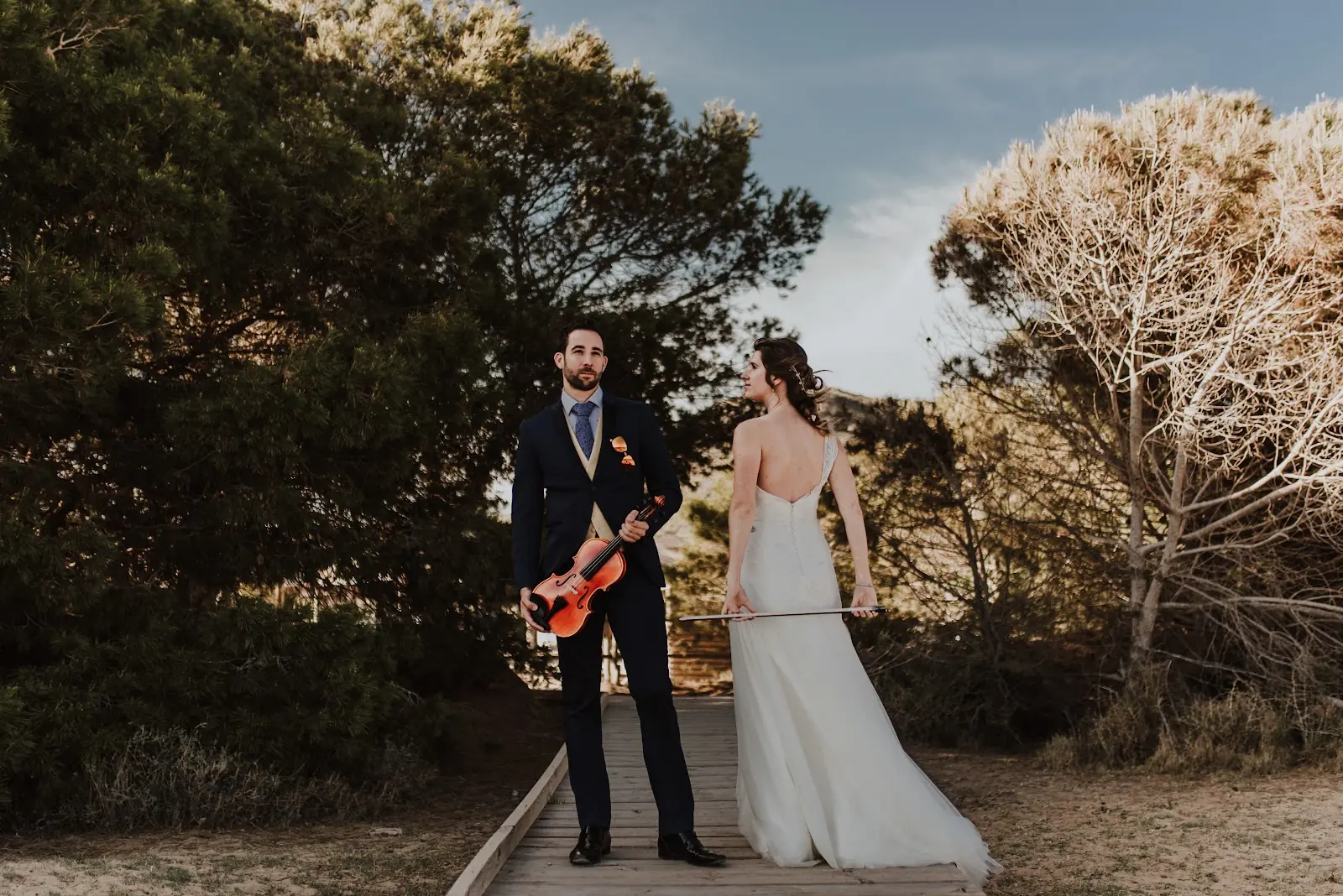 Bride and groom posing with a violin on a wooden path among pine trees, photograph by José Espinosa