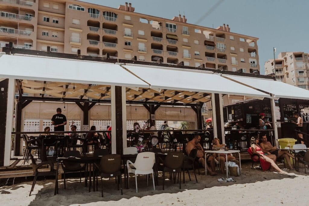 Chiringuito A Tu Aire en La Manga del Mar Menor con terraza bajo toldo blanco llena de clientes en bañador y barra de bar al fondo