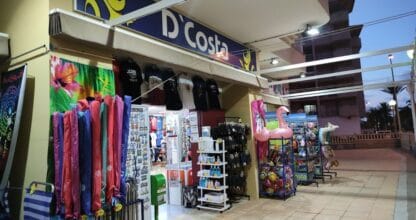 Tienda D'Costa en La Manga del Mar Menor con artículos de playa, toallas, flotadores y souvenirs expuestos en la entrada al atardecer