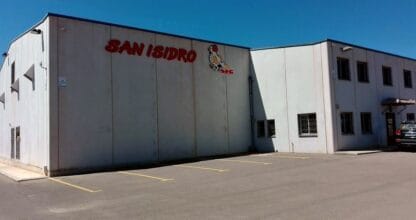 Nave industrial de Avícola San Isidro con letrero rojo y logo de gallina, aparcamiento exterior bajo cielo azul