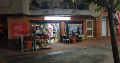 Fachada nocturna del Bazar LAI con macetas, plantas y lista de productos en pared roja, La Manga del Mar Menor