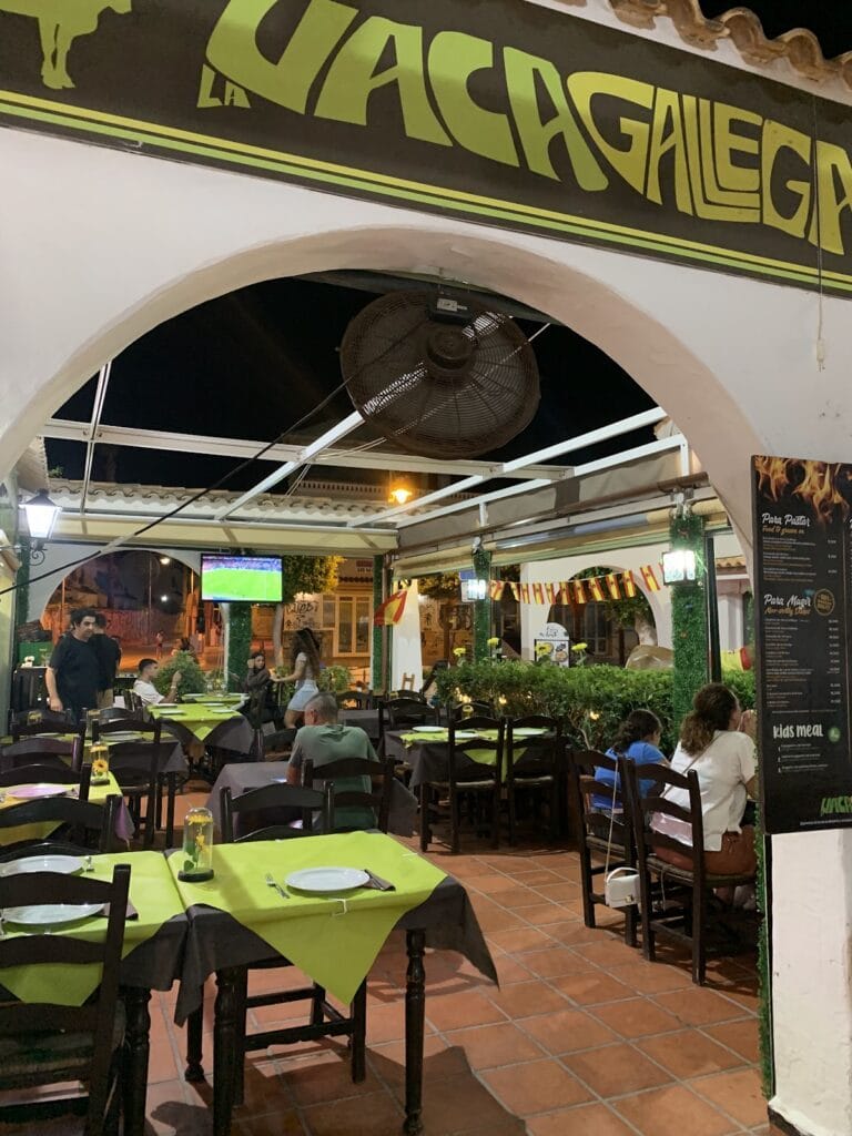 Terraza nocturna de Brasería La Vaca Gallega con mesas verdes, clientes cenando y televisión en La Manga