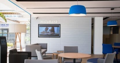 Sala de espera de CaixaBank HolaBank con sillones grises, lámparas azules y pantalla corporativa en ambiente moderno