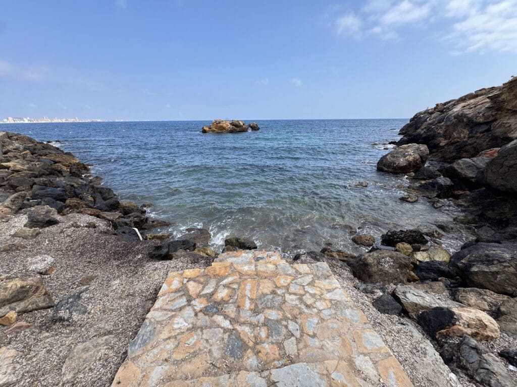 Cala rocosa con aguas azules y transparentes y acceso de piedra en Cala de la Galera, Cabo de Palos