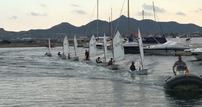 Escuela de vela del Club Náutico La Isleta con optimists navegando en fila acompañados por zodiac, montañas al fondo en La Manga del Mar Menor