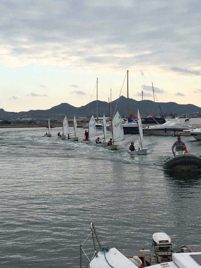 Escuela de vela del Club Náutico La Isleta con optimists navegando en fila acompañados por zodiac, montañas al fondo en La Manga del Mar Menor