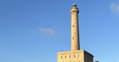 Faro histórico de Cabo de Palos sobre colina rocosa con matorral mediterráneo bajo cielo azul, zona de la Cruz Memorial Víctimas del Sirio