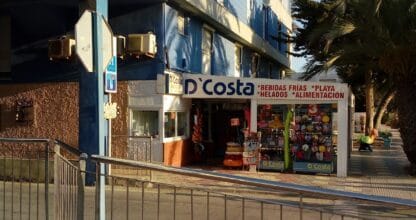 Fachada de D'Costa con rótulo de bebidas frías, helados y artículos de playa junto a palmeras en La Manga del Mar Menor