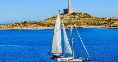 Velero de Djinn Charter navegando con velas desplegadas frente al faro de Cabo de Palos en aguas azules del Mediterráneo