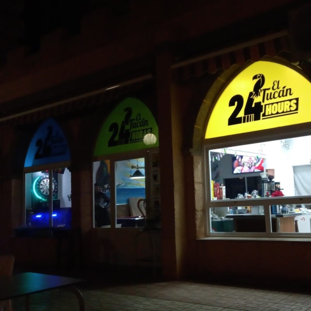 Fachada nocturna de El Tucan 24 Hours en La Manga del Mar Menor con rótulos luminosos de colores y diana de dardos al fondo