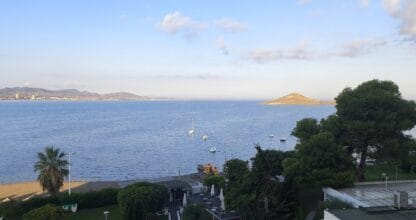 Vista del Mar Menor desde un edificio en La Manga con veleros fondeados, playa arbolada y montículo rocoso al fondo al atardecer