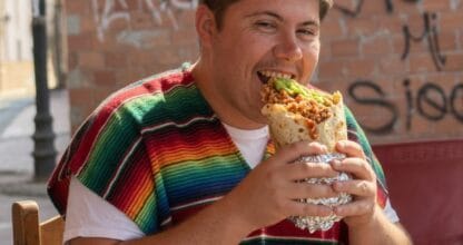 Joven con poncho mexicano de colores mordiendo un enorme burrito en terraza exterior de FanatiCamis