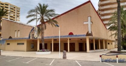 Iglesia de Santa María Reina de los Apóstoles con palmeras y mosaico en fachada entre edificios de La Manga del Mar Menor