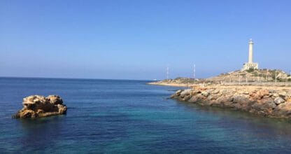 Vista del faro de Cabo de Palos desde el mar con aguas cristalinas azul turquesa y rocas, imagen de la zona que cubre Inmobiliaria Abacasa