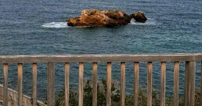 Islote rocoso de la Isla del Pato Vizo visto desde una barandilla de madera sobre el Mediterráneo, con volcán al fondo en el horizonte