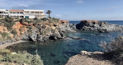 Pequeña cala de la Isla Poncho con agua azul turquesa entre acantilados rojizos y edificio blanco sobre el promontorio en Cabo de Palos
