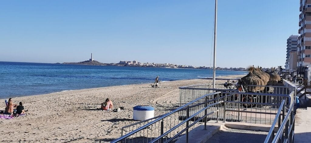Vista de la playa de La Manga del Mar Menor desde Jijonenca Helado Artesano con bañistas en la arena y el faro de Cabo de Palos al fondo