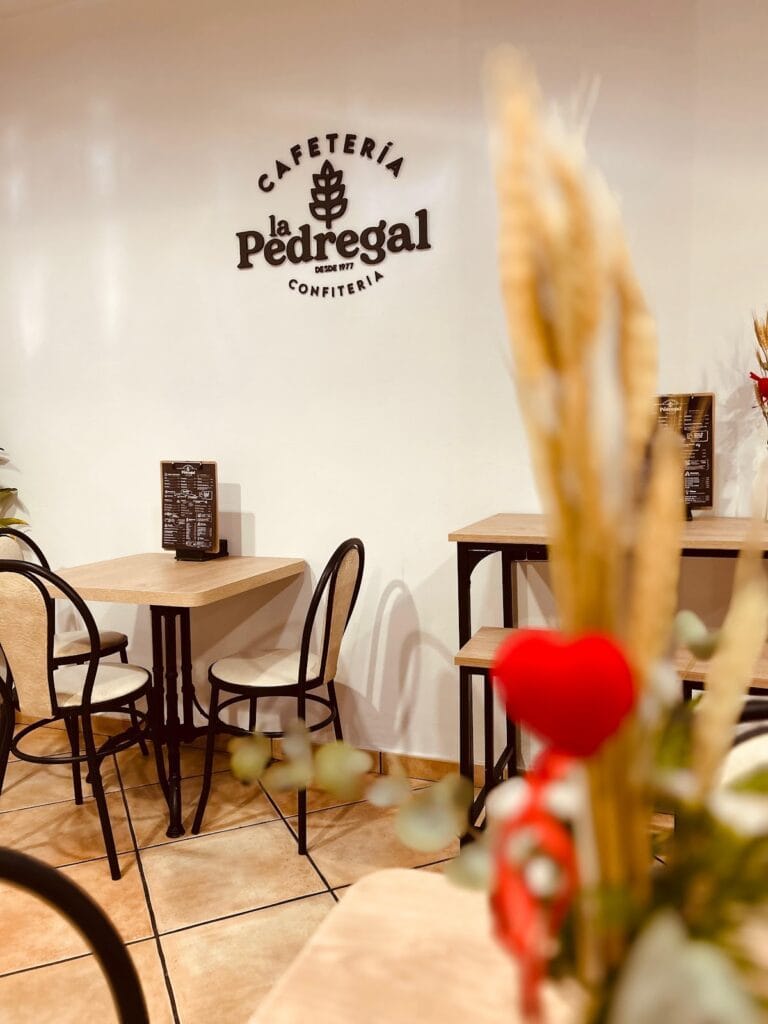 Interior de Cafetería La Pedregal desde 1977 con logo en pared blanca, mesas de madera y sillas negras