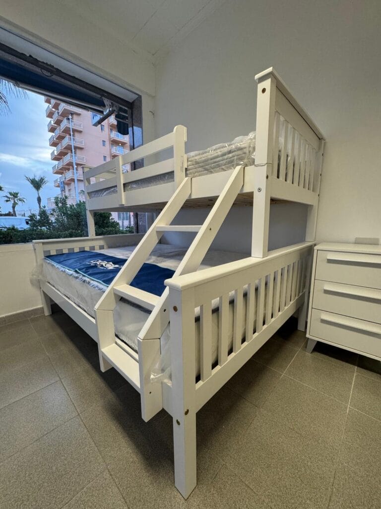 Litera de madera blanca de dos pisos en exposición de Madera y Mar en La Manga del Mar Menor, con vistas a edificios desde la ventana del local