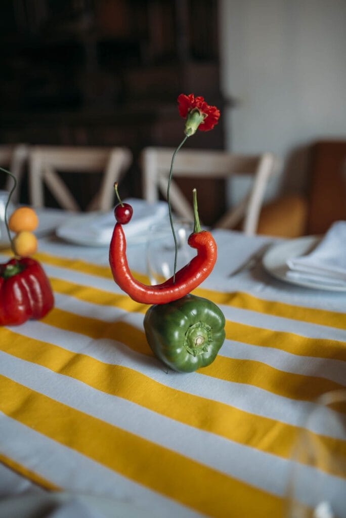 Decoración de mesa creativa en MaracaStudio con pimiento verde, guindilla roja y clavel formando una composición artística sobre mantel de rayas amarillas