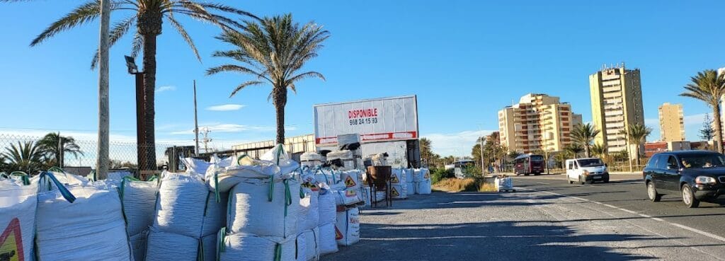 Almacén exterior de materiales de construcción MANGAMAT con sacos apilados junto a palmeras y edificios en La Manga del Mar Menor