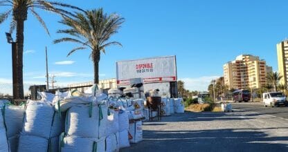 Almacén exterior de materiales de construcción MANGAMAT con sacos apilados junto a palmeras y edificios en La Manga del Mar Menor