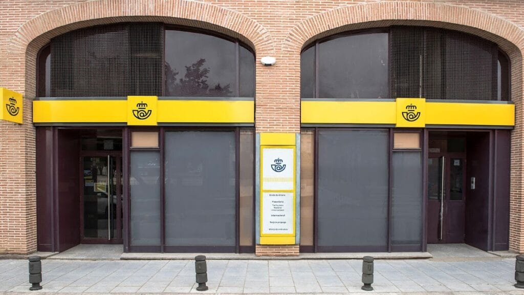 Fachada de oficina de Correos con rótulos amarillos y logo corporativo en edificio de ladrillo visto con doble acceso