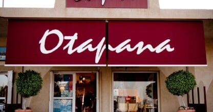 Fachada de la tienda Otapana con gran toldo rojo, escaparates y árboles en macetas, La Manga del Mar Menor