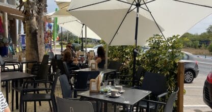 Terraza exterior de la Panadería Cafetería Pura Vida en La Manga del Mar Menor con mesas negras, sombrillas blancas y palmeras