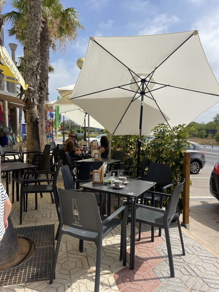 Terraza exterior de la Panadería Cafetería Pura Vida en La Manga del Mar Menor con mesas negras, sombrillas blancas y palmeras