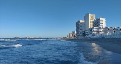 Playa las Sirenas en La Manga del Mar Menor con olas rompiendo en la orilla arenosa, edificios de apartamentos y faro al fondo al atardecer
