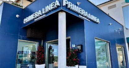 Fachada azul de Primera Línea Inmobiliarias La Manga con grandes rótulos blancos, flores rojas y escaparates de propiedades
