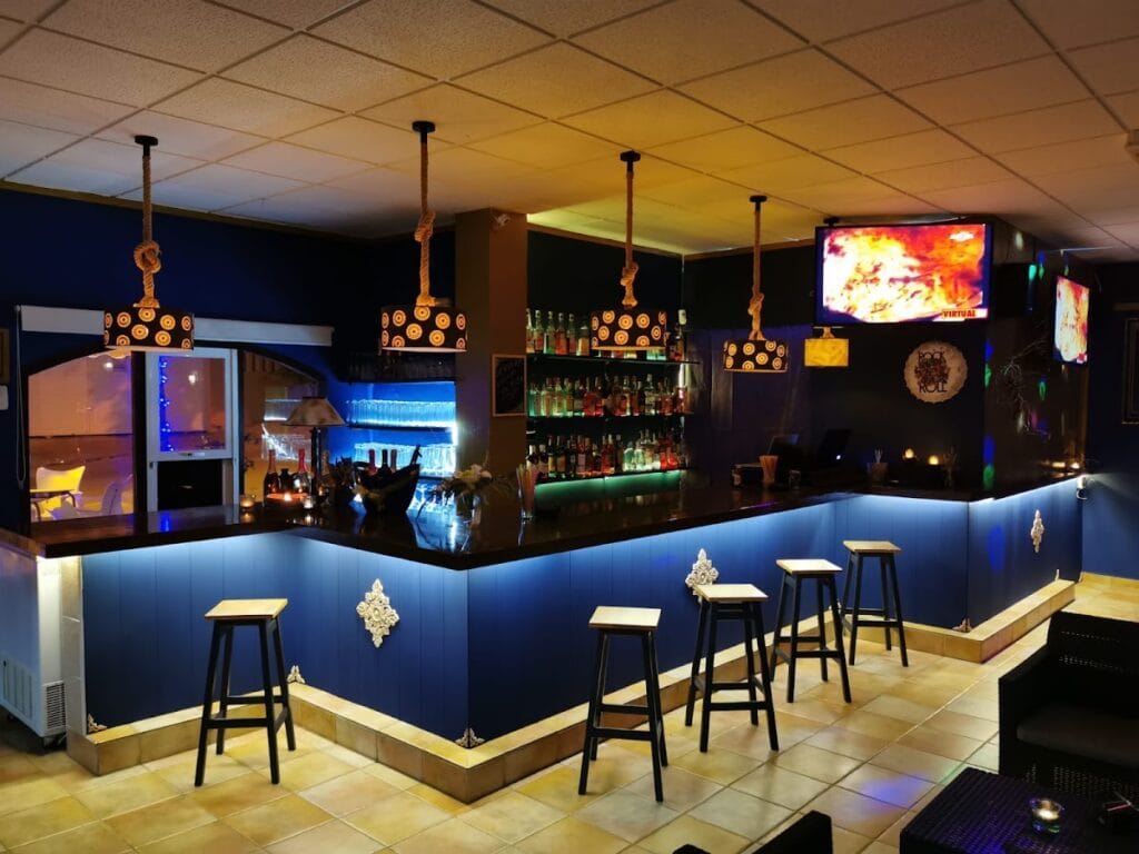 Barra de cócteles iluminada en azul con taburetes altos y pantalla de televisión en el Pub Emporio Gourmet y Copas