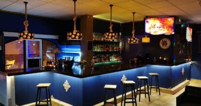 Barra de cócteles iluminada en azul con taburetes altos y pantalla de televisión en el Pub Emporio Gourmet y Copas