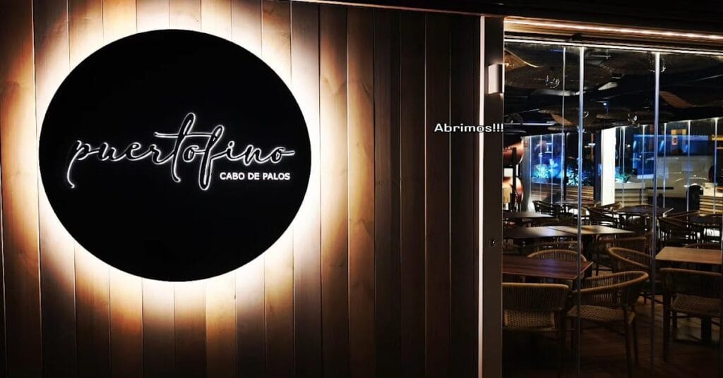 Entrada nocturna de Puertofino Cabo de Palos con logo circular iluminado en madera y sala de comedor moderna al fondo