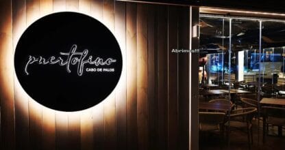 Entrada nocturna de Puertofino Cabo de Palos con logo circular iluminado en madera y sala de comedor moderna al fondo