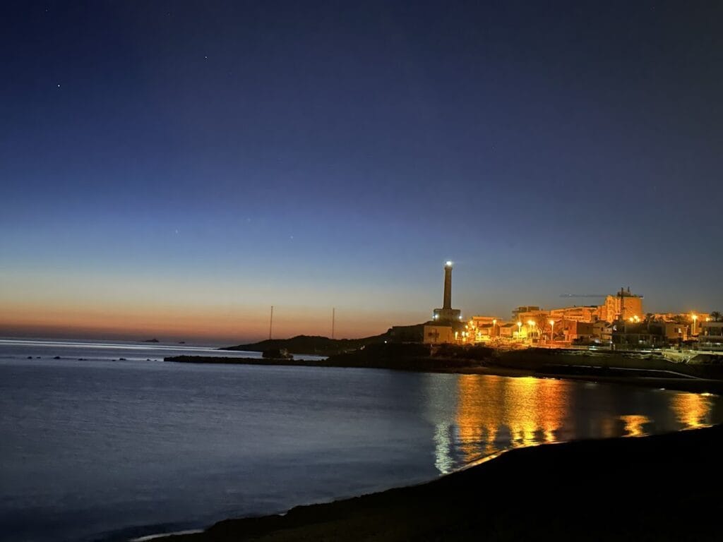 Vista nocturna del faro de Cabo de Palos iluminado junto al pueblo marinero y su reflejo dorado en el agua tranquila