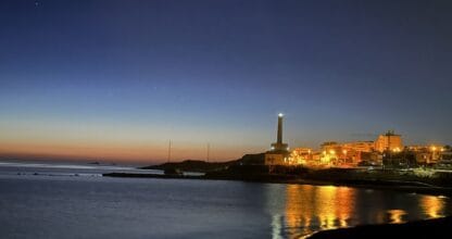 Vista nocturna del faro de Cabo de Palos iluminado junto al pueblo marinero y su reflejo dorado en el agua tranquila