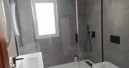 Baño reformado con plato de ducha blanco, mampara negra y revestimiento gris, trabajo de Reformas Mangadecor
