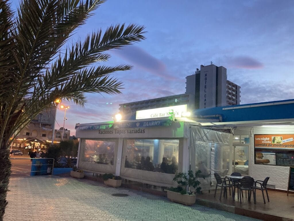 Exterior nocturno del Café Bar Nino's en La Manga del Mar Menor, con terraza acristalada y letrero de raciones y tapas variadas