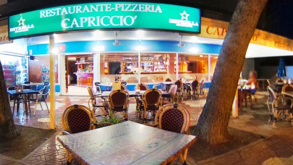 Fachada nocturna del Restaurante-Pizzería Capriccio del Perlita con gran rótulo verde iluminado y terraza con sillas de mimbre