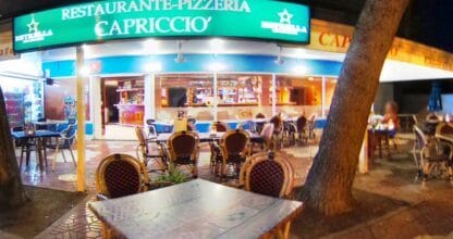 Fachada nocturna del Restaurante-Pizzería Capriccio del Perlita con gran rótulo verde iluminado y terraza con sillas de mimbre