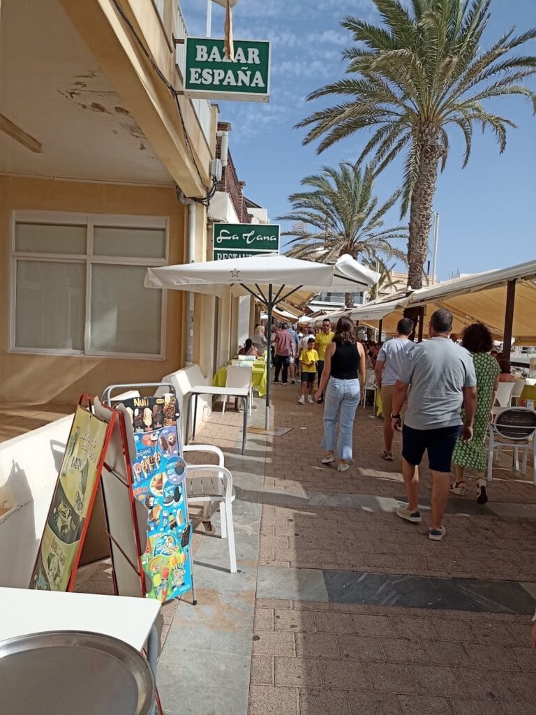 Calle peatonal animada de La Manga del Mar Menor con terrazas de bares, palmeras y turistas paseando junto a Spar Express
