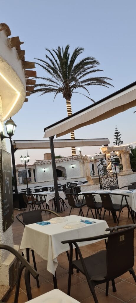 Terraza exterior de St. Gregory's Restaurante A La Brasa al atardecer, con mesas de mantel blanco, palmera y jardín porticado