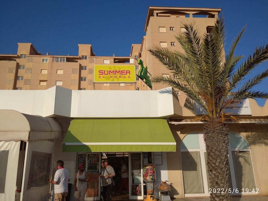 Fachada del comercio Summer 24 Horas en La Manga del Mar Menor con toldo verde, palmera y clientes en la entrada al atardecer