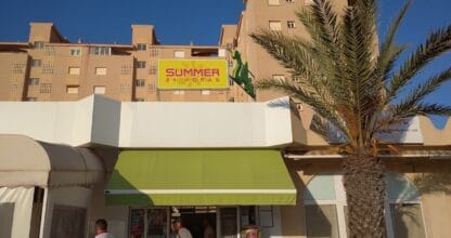 Fachada del comercio Summer 24 Horas en La Manga del Mar Menor con toldo verde, palmera y clientes en la entrada al atardecer